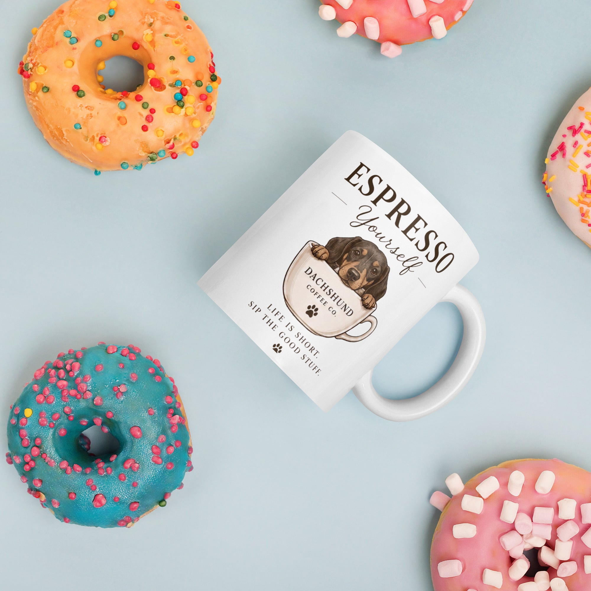 Espresso Yourself Dachshund Coffee Cup laying on a blue background surrounded by colourful donuts